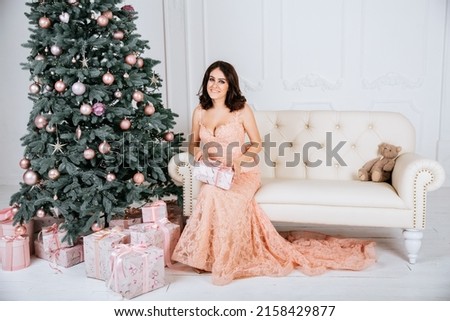 Brunette woman in orange dress smiling near Christmas tree sitting on sofa with present (authoring photo)