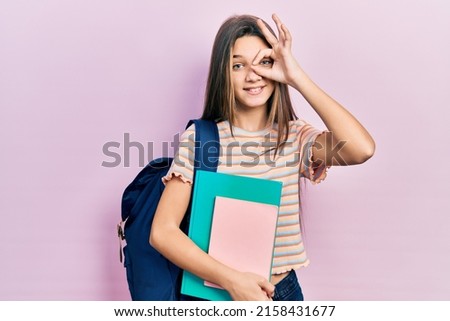 Young brunette girl holding student backpack and books smiling happy doing ok sign with hand on eye looking through fingers 