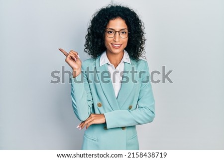Young latin girl wearing business clothes and glasses with a big smile on face, pointing with hand and finger to the side looking at the camera. 