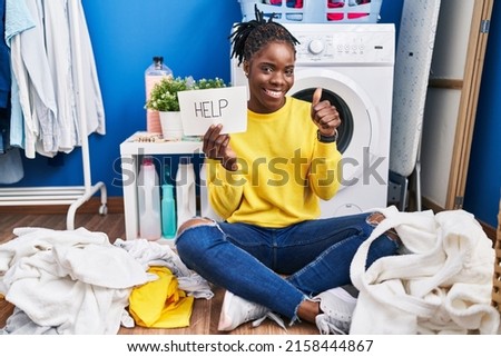 Beautiful black woman doing laundry asking for help smiling happy and positive, thumb up doing excellent and approval sign 