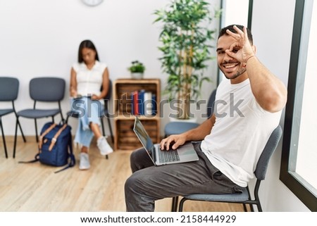 Young people sitting at waiting room working with laptop smiling happy doing ok sign with hand on eye looking through fingers 