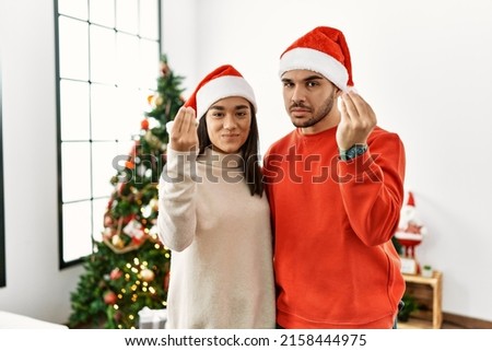 Young hispanic couple standing by christmas tree doing italian gesture with hand and fingers confident expression 