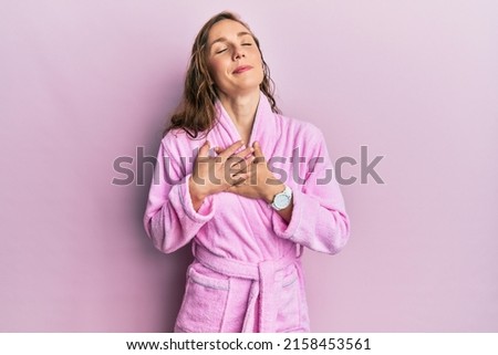 Young blonde woman wearing bathrobe smiling with hands on chest with closed eyes and grateful gesture on face. health concept. 