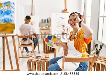 Young artist woman painting on canvas at art studio doing ok gesture with hand smiling, eye looking through fingers with happy face. 