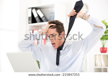 Stressed young Businessman sitting in the office. He is angry holding necktie and looking in laptop.