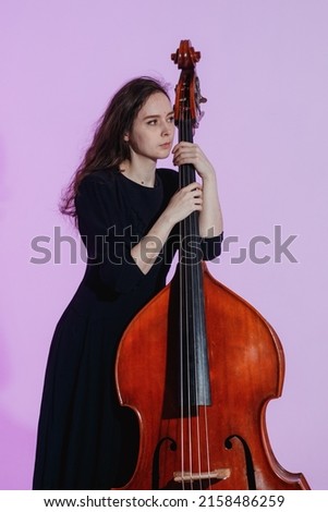 Girl musician with long hair in black dress poses with the double bass against the light purple background.