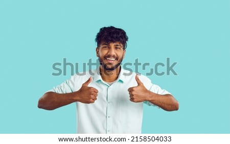 Studio shot of happy attractive bearded mustached young Indian man with curly black hair in smart casual white shirt standing on turquoise background, giving thumbs up, looking at camera and smiling