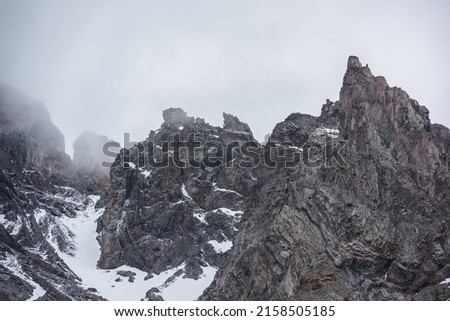 Gloomy mountain landscape with high sharp rockies with snow in gray low clouds. Closeup of sharp rocks and peaked tops in gray cloudy sky. Bleak overcast scenery with mountain range with pointy rocks. Royalty-Free Stock Photo #2158505185