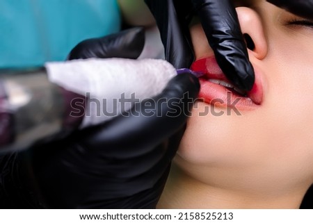 the tattoo artist holds the lips with his fingers, thereby turning them and shading the lower contour of the lips with red pigment using a tattoo machine Royalty-Free Stock Photo #2158525213