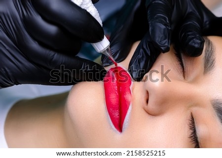 the master performs the procedure of permanent makeup of the lips holding them while applying a tattoo machine for tattoo Royalty-Free Stock Photo #2158525215