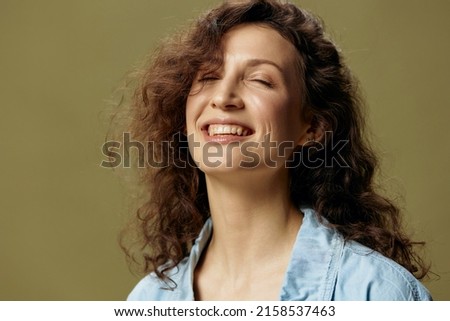 Cheerful happy curly beautiful female in jeans casual shirt smiles closing eyes posing isolated on over olive green pastel background. Being Yourself. People Lifestyle emotions concept. Copy space