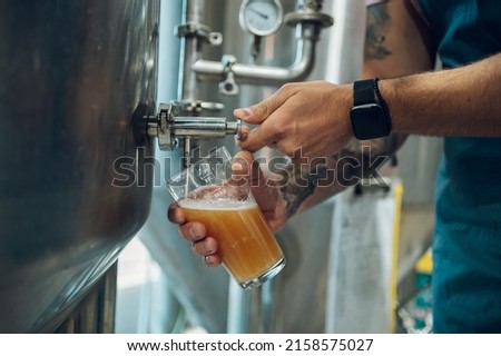 Man examining the quality of craft beer at brewery. Male brewer working at beer factory checking the beer quality. Testing beer at a modern brewery. Cropped shot of a man filling glass. Royalty-Free Stock Photo #2158575027