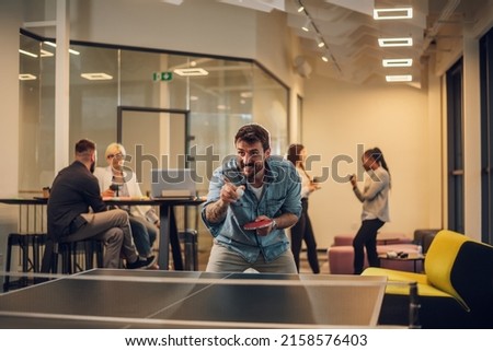 Young people playing table tennis in workplace and having fun. Concept of sport, friendship, teambuilding and teamwork. Focus on a business man having fun while his colleagues having a meeting .