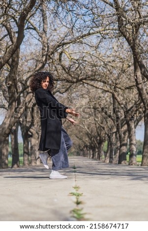 A curly Spanish woman dancing on the road against bare trees on a sunny day