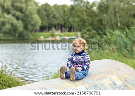 A little girl toddler is sitting on the shore of a lake in the spring forest. Nature walk.