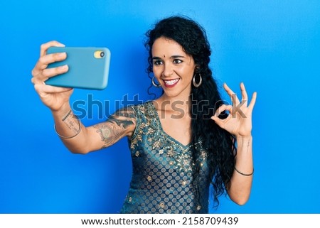 Young woman wearing bindi and traditional kurta dress taking a selfie photo with smartphone doing ok sign with fingers, smiling friendly gesturing excellent symbol 