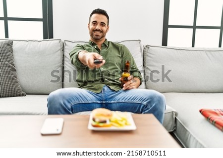 Young hispanic man eating hamburger watching tv at home
