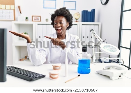 African dentist woman working at medical clinic amazed and smiling to the camera while presenting with hand and pointing with finger. 