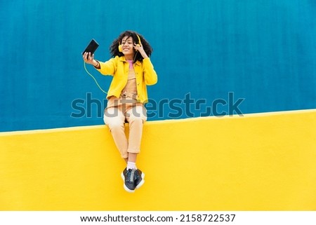 Young happy woman dancing and having fun outdoor. Teenager listening to music with smartphone and headphones in a yellow and blue modern urban area Royalty-Free Stock Photo #2158722537