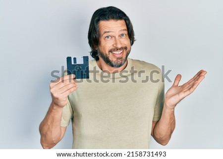 Middle age caucasian man holding floppy disk celebrating achievement with happy smile and winner expression with raised hand 