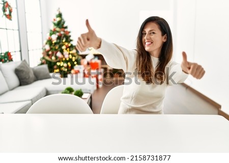 Beautiful caucasian woman sitting on the table by christmas tree approving doing positive gesture with hand, thumbs up smiling and happy for success. winner gesture. 
