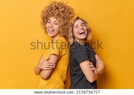 Positive two young pretty women stand back to each other keep arms folded laugh happily dressed in casual t shirts laugh at something funny isolated over yellow background. Friendship concept Royalty-Free Stock Photo #2158736757