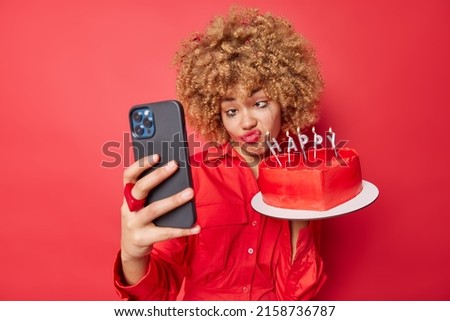 Funny curly haired young woman makes grimace holds tasty heart cake wears shirt takes selfie via smartphone foolishes around prepares for birthday celebration. Drunk female on party with dessert