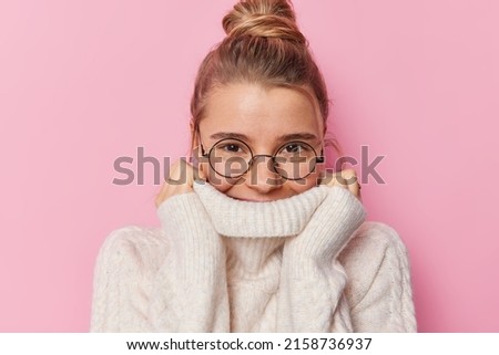 Beautiful young woman with fair hair gathered in bun hides mouth behind collar of sweater wears round spectacles isolated over pink background. Sincere delighted female model wears warm jumper Royalty-Free Stock Photo #2158736937