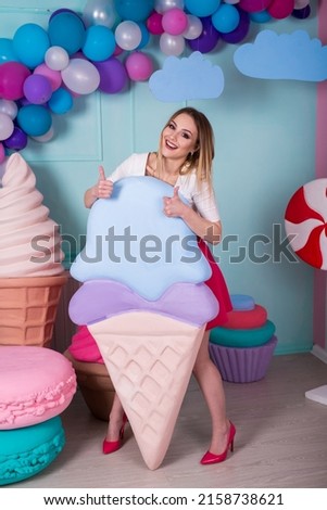 Portrait of young woman in pink dress holding big ice cream and posing on decorated background. Amazing sweet-tooth girl surrounded by toy sweets. 
