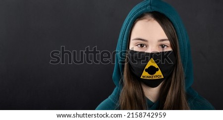 MONKEYPOX. Teenage girl in a medical mask. A sign with a danger sign and a monkey on the mask. Virus, epidemic, disease.Black background. Royalty-Free Stock Photo #2158742959