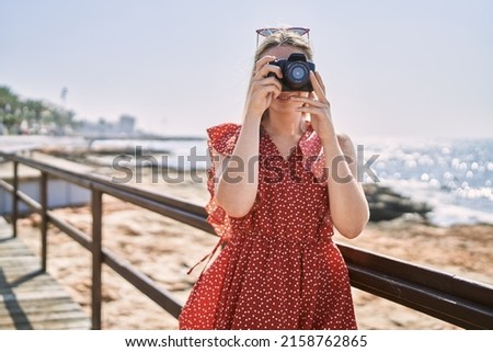 Young blonde girl smiling happy using camera at the beach