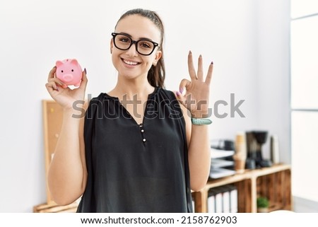 Young brunette woman holding piggy bank at the office doing ok sign with fingers, smiling friendly gesturing excellent symbol  Royalty-Free Stock Photo #2158762903