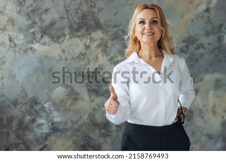 Smiling older mature businesswoman employer showing thumbs up like hand gesture standing isolated on textured background, banner, copy space.