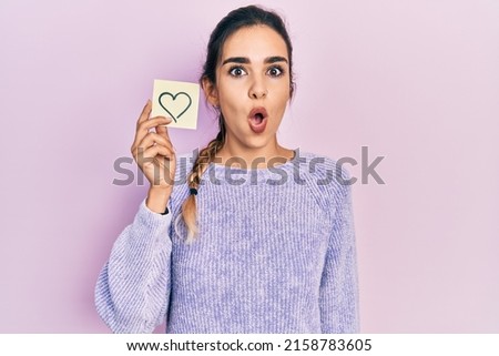 Young hispanic girl holding heart reminder scared and amazed with open mouth for surprise, disbelief face 