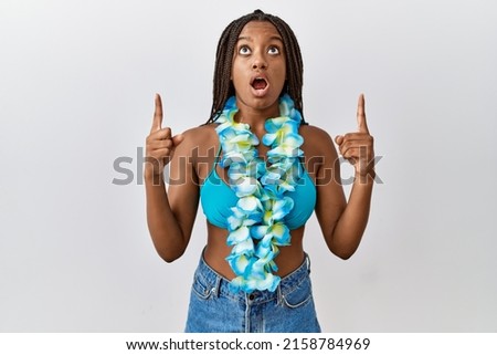 Young african american woman with braids wearing bikini and hawaiian lei amazed and surprised looking up and pointing with fingers and raised arms. 