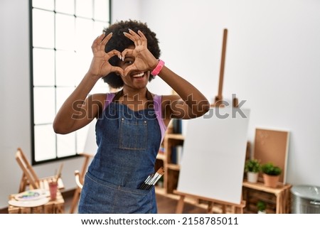 Young african american woman with afro hair at art studio doing heart shape with hand and fingers smiling looking through sign 