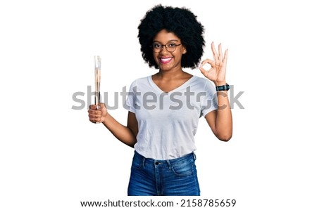 Young african american woman holding paintbrushes doing ok sign with fingers, smiling friendly gesturing excellent symbol 