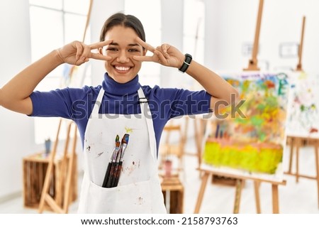 Young brunette woman at art studio doing peace symbol with fingers over face, smiling cheerful showing victory 