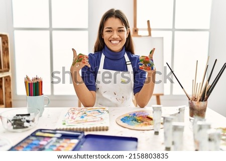 Young brunette woman at art studio with painted hands success sign doing positive gesture with hand, thumbs up smiling and happy. cheerful expression and winner gesture. 