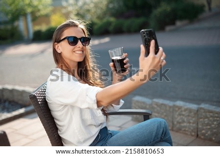 Positive young woman taking selfie on modern smartphone while resting on outdoors terrace of hotel. Happy female brunette in sunglasses an casual wear holding glass of refreshing drink.