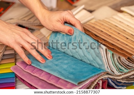 Fabric swatches in different colors are stacked for selection. A woman chooses upholstery colors for furniture and interiors. Touches the texture of the fabric, hands on the material close-up.