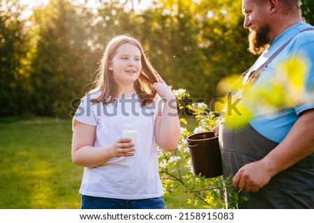 Happy teenager girl drinks goat milk outdoor from a glass and her father with love looks at her.