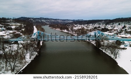An aerial beautiful view of large metal blue bridge over a river water with snowy trees and a gray sky
