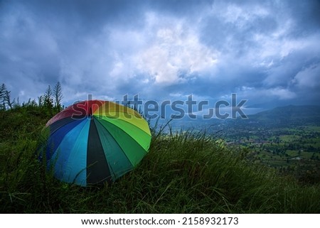 A rainbow colored umbrella enjoying and relaxing on the green hillside against the dramatic blue backdrop of monsoon clouds, beautifully lit green fields and the glistening waters of a lake.    Royalty-Free Stock Photo #2158932173
