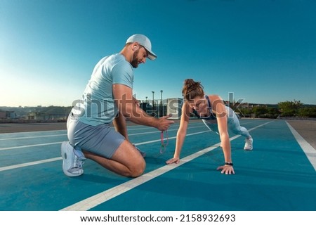 Instructor and athlete runner on the track. The athletics marks the time in the plank exercise on a stopwatch. Fitness trainer and mentee. Royalty-Free Stock Photo #2158932693