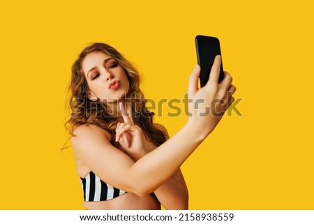 portrait of beautiful young woman texting flirt selfie photo message with smartphone on yellow background studio