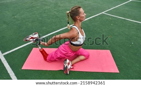 Side view of athletic girl with artificial leg has disability warming up on fitness mat on sports field. Young focused caucasian woman wear sportswear. Healthy lifestyle. Conquering adversity Royalty-Free Stock Photo #2158942361