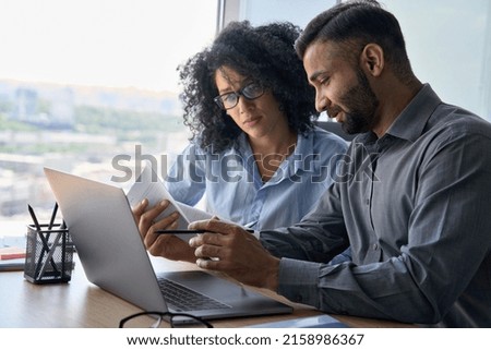 Multiethnic male indian mentor and female African American intern sitting at desk with laptop doing paperwork together discussing project financial report. Corporate business collaboration concept. Royalty-Free Stock Photo #2158986367