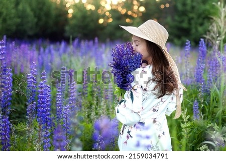 View from the back of a young girl in a dress and hat with a bouquet of lupins in her hands on a background of green meadow. Vertical
