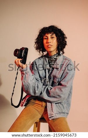 Confident young photographer looking at the camera while holding a digital camera. Freelance photographer sitting on a chair against a studio background.